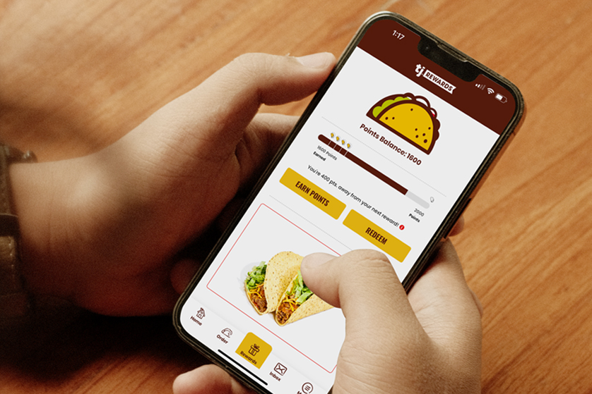 Hand holding a smartphone displaying the Taco John’s rewards app with points balance, “Earn Points” and “Redeem” buttons, and an image of tacos on screen.