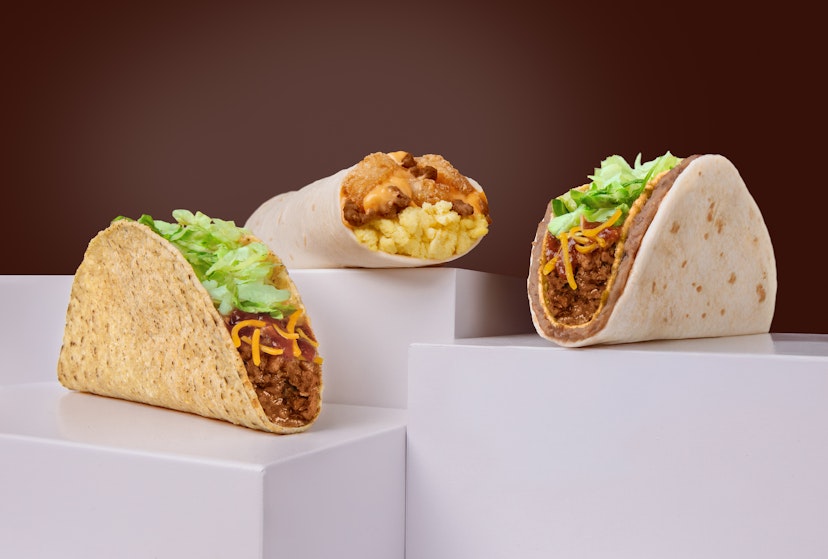 Three Taco John’s daily deal items on white pedestal blocks: Crispy Taco with beef, lettuce, and cheese; Taco Bravo with layers of meat and toppings in a soft tortilla; and a Meat & Potato breakfast burrito filled with eggs, potatoes, and meat.